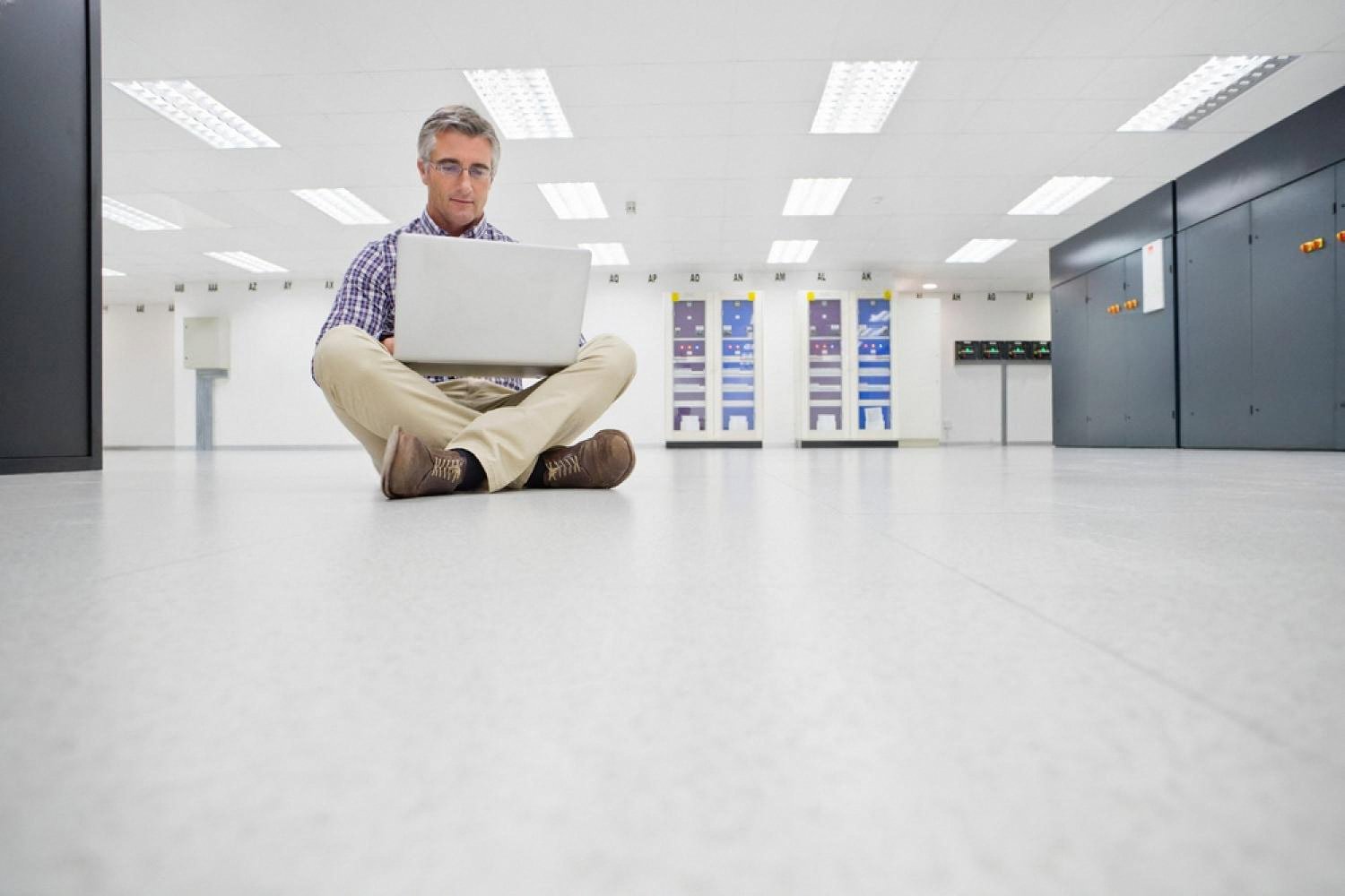 Man sitting on the floor of a data centre with a laptop, reviewing hosting configuration